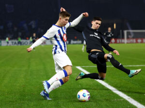 Gustav Christensen lægger an til at sparke til en bold, mens en forsvarsspiller kaster sig ned for at blokere.