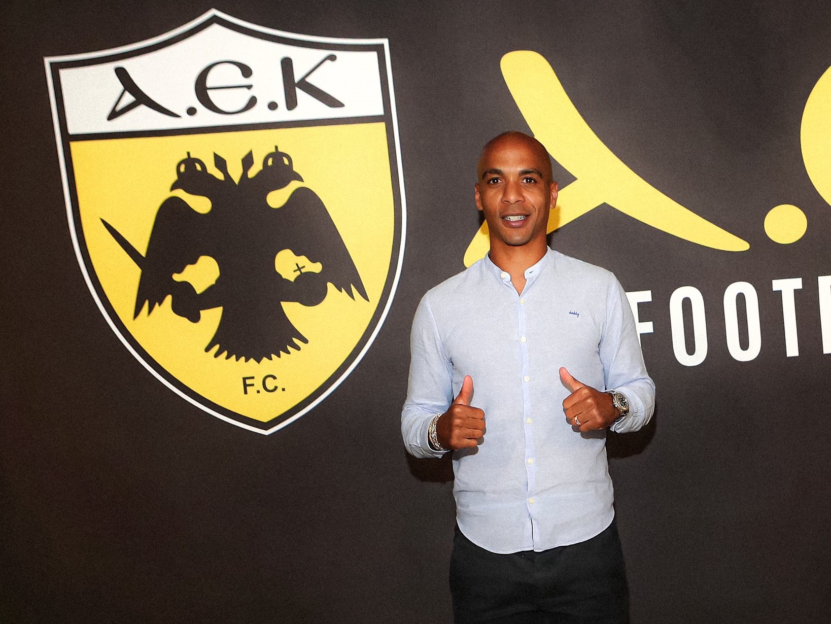 Portugisiske Joao Mario med to tommefingre op og et AEK logo i baggrunden.