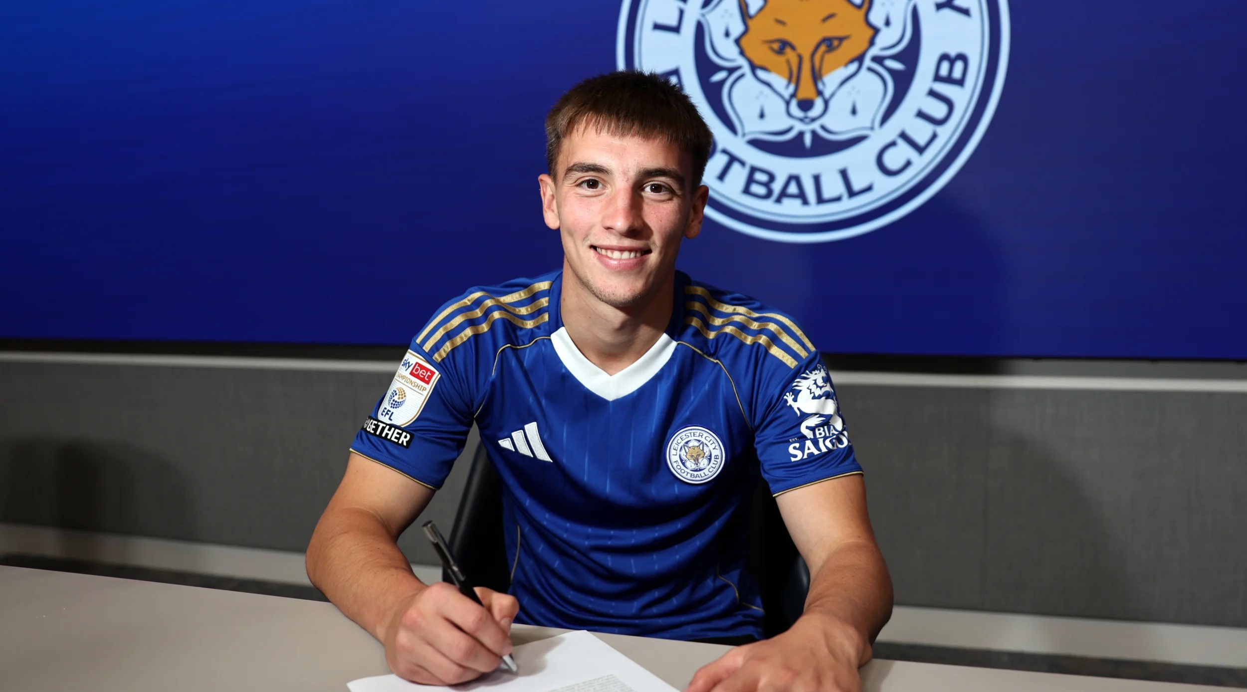 Luke Thomas skriver under på en kontrakt med et Leicester-logo i baggrunden.
