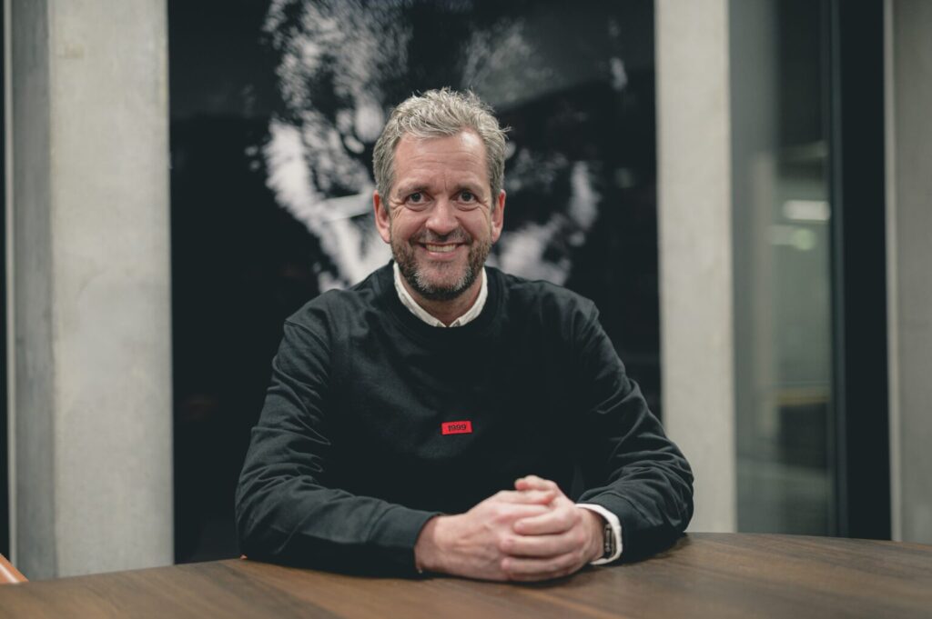 Kristian kamp sidder med foldede hænder ved et bord.