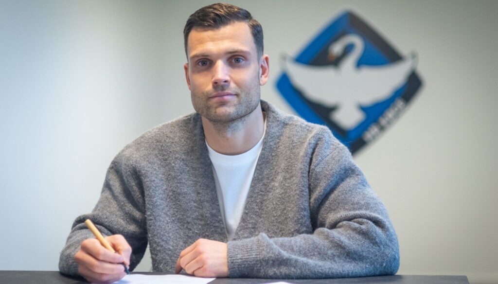 Tobias Thomsen skriver under på en kontrakt med et HB Køge logo i baggrunden.